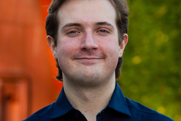 A young man with light brown hair and a slight smile stands outdoors, wearing a dark blue shirt. The background features greenery and a blurred structure